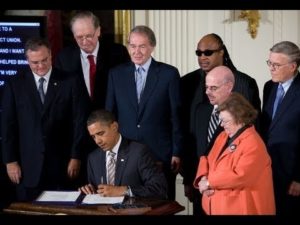 President Obama signs a law while sitting at desk.  He is surrounded by others including Senators and Stevie Wonder.